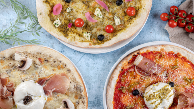 Trois pizzas artisanales présentées sur fond bleu clair : l’une au pesto, tomates cerises et feta, l’autre à la truffe et burrata, la dernière avec jambon cru et mozzarella